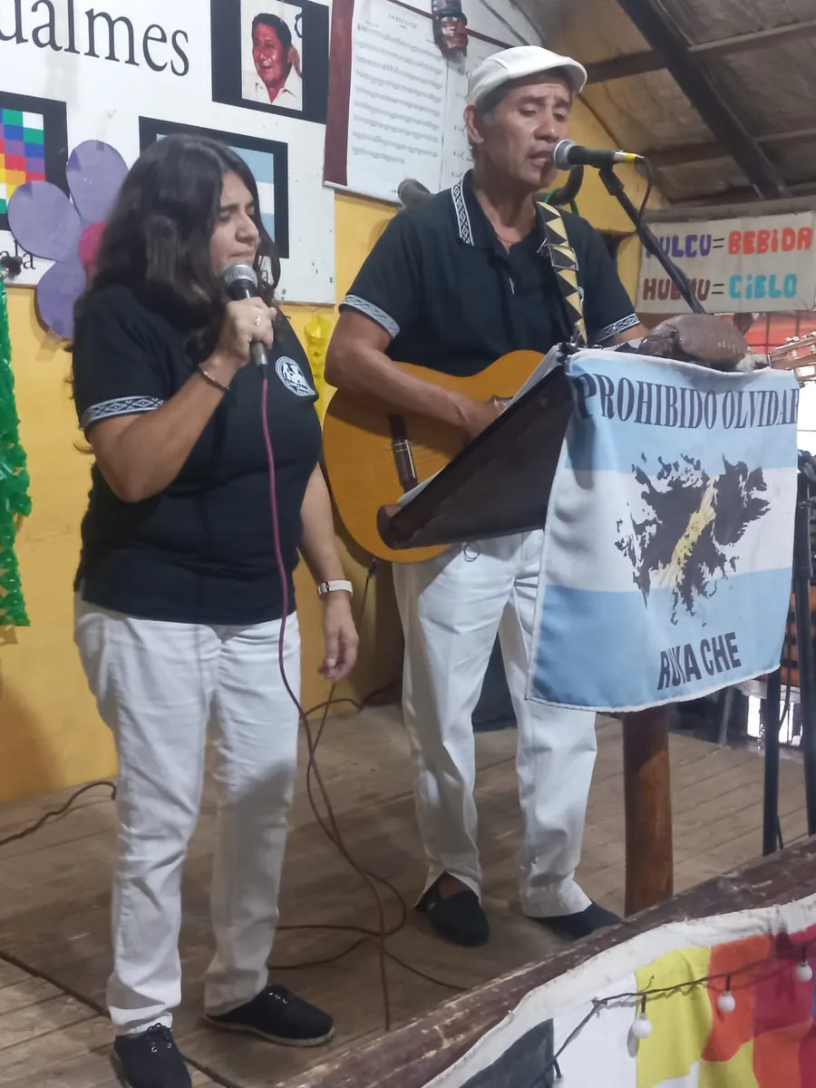 El dúo cantando en un escenario comunitario junto a la bandera de Malvinas